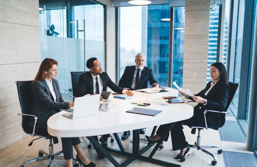 Four colleagues at an oval meeting table, with towers beyond the window